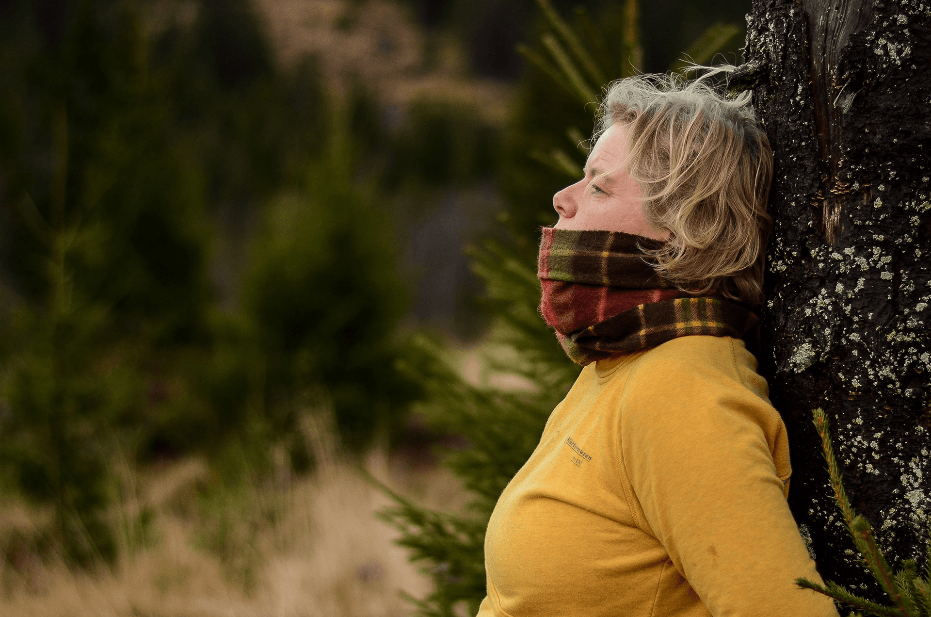 A woman in profile with her eyes closed, leaning against a tree trunk in a forest at golden hour, wearing a yellow jumper and a plaid scarf.