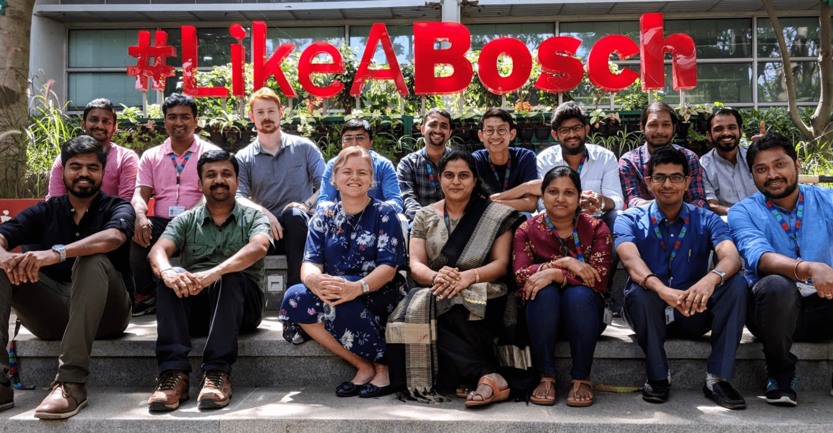 A large group of Bosch India colleagues sitting together outdoors in front of a large red "#LikeABosch" sign.