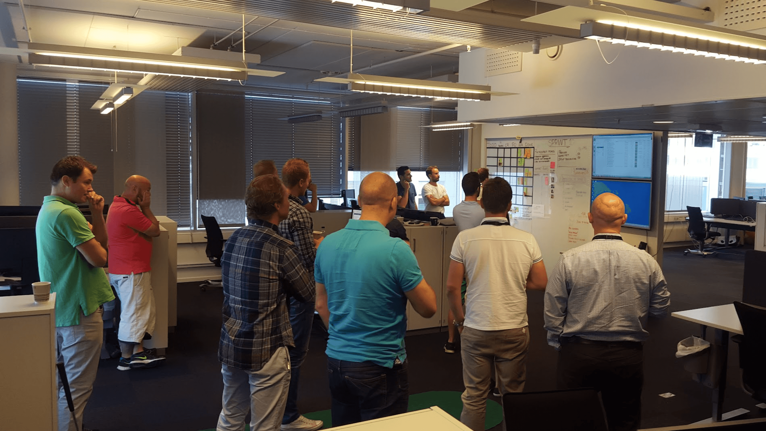 A group of around ten colleagues standing together reviewing a sprint board and screens in an open office.
